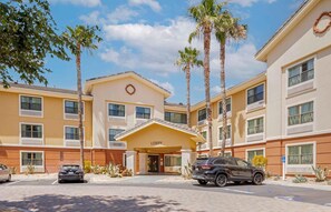A hotel with a yellow facade, a 'Lobby' sign, palm trees, and a parking lot with cars.