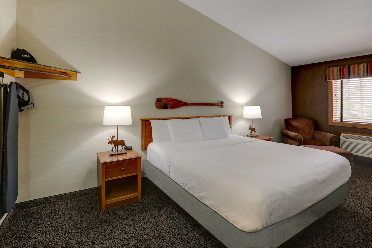 A hotel room with a large bed, two bedside lamps, a wooden nightstand, a chair, and a window with blinds.