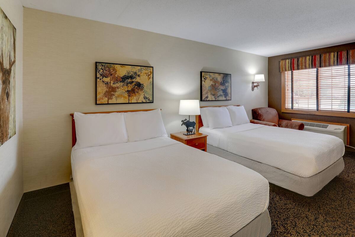 A hotel room with two beds, a nightstand with a lamp, a decorative cow figurine, and artwork on the walls.