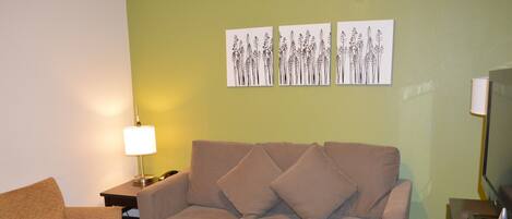 A living room with a sofa, armchair, and coffee table. There is a lamp, a TV, and artwork on the wall.