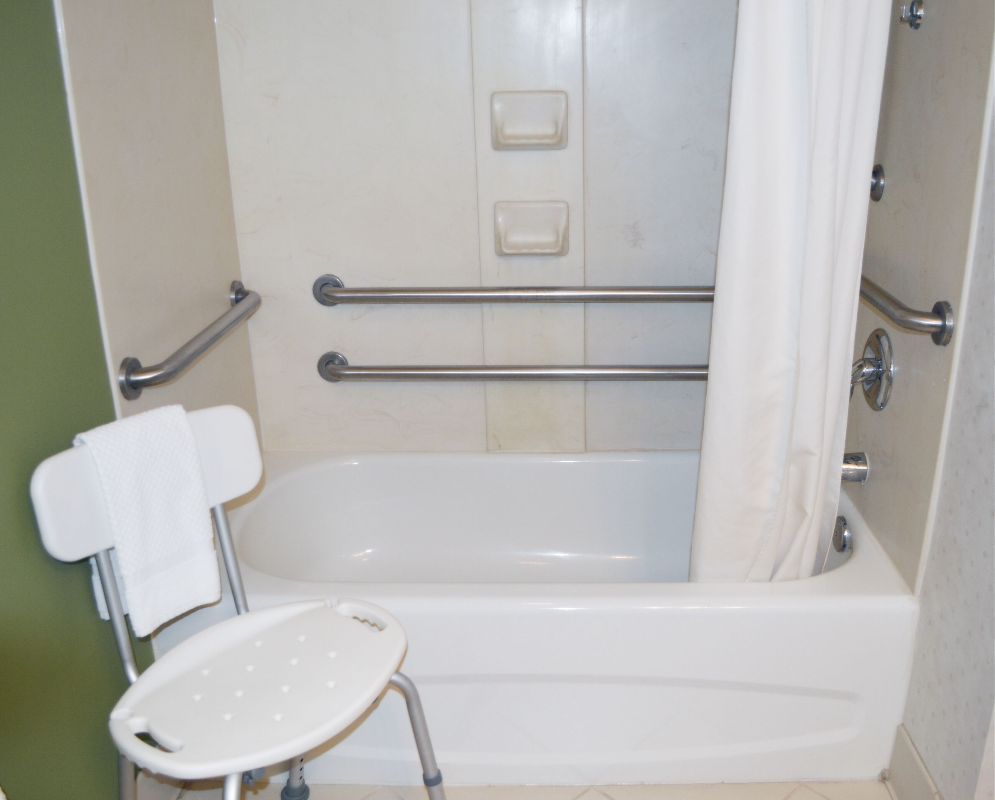 Une salle de bain avec une baignoire, des barres d’appui, un rideau de douche et une chaise blanche.