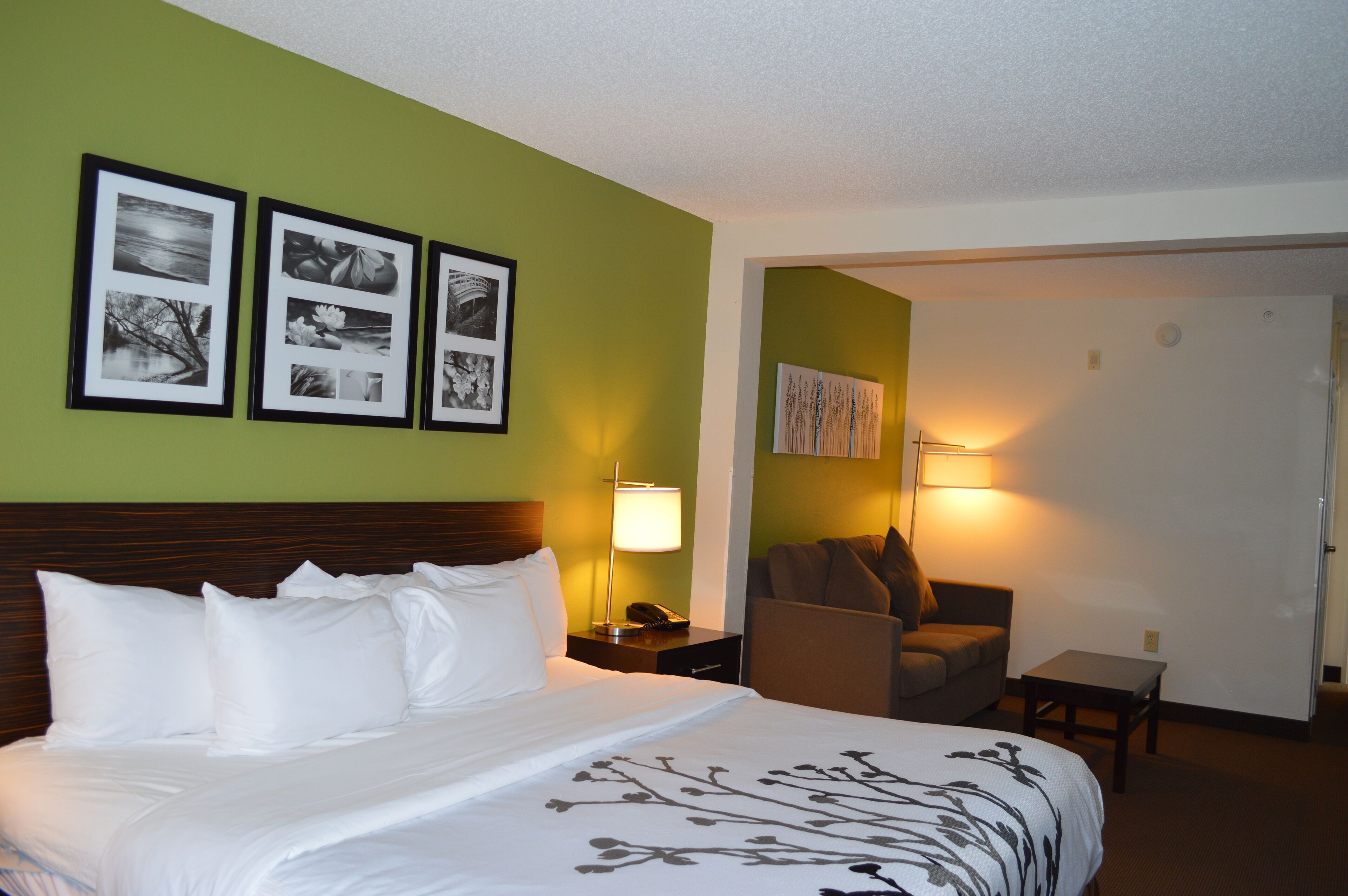 A hotel room with a large bed, a nightstand, a lamp, a chair, and a small table.