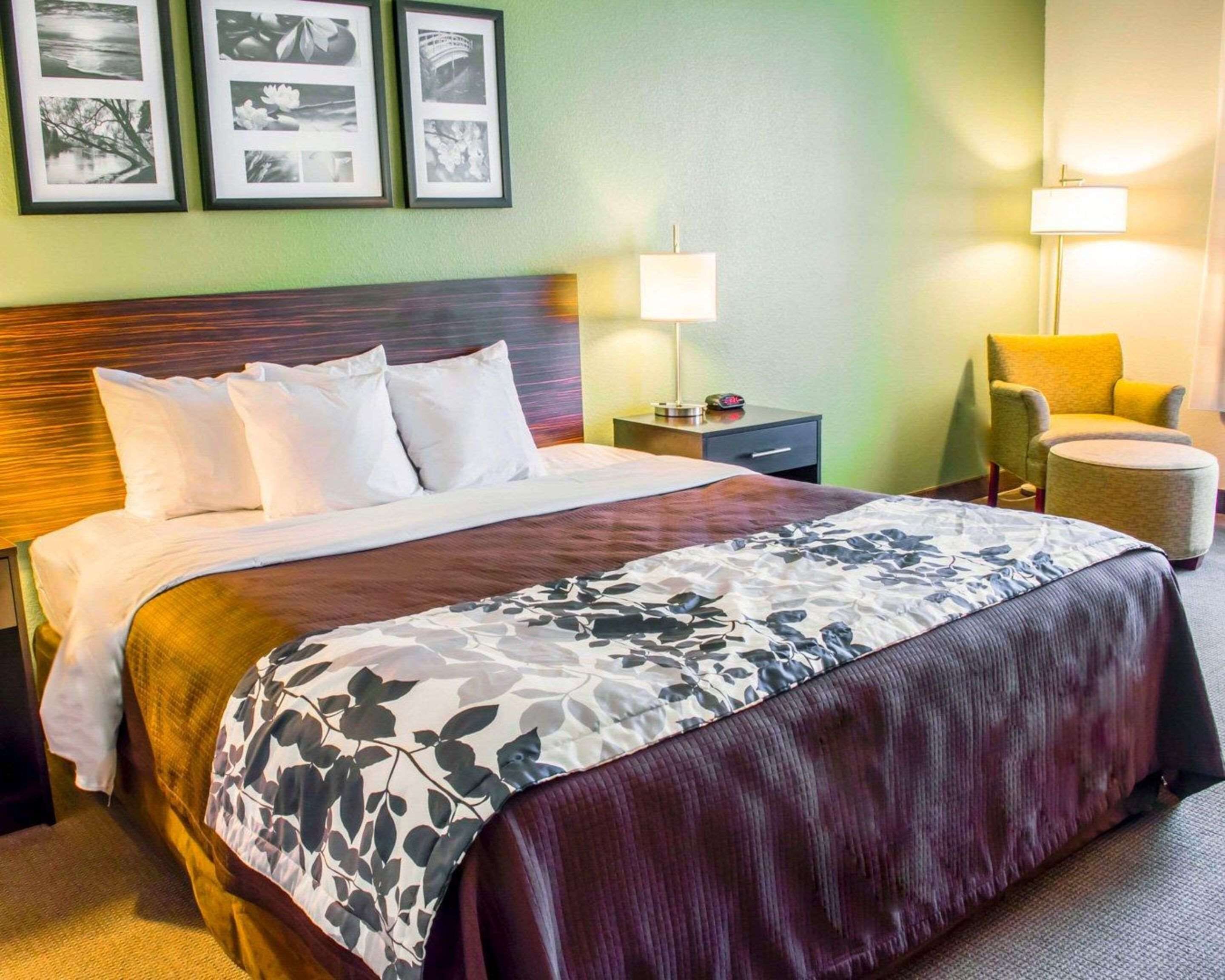 Una habitación de hotel con una cama grande, una mesita de noche con lámpara, una silla y cuadros en la pared.
