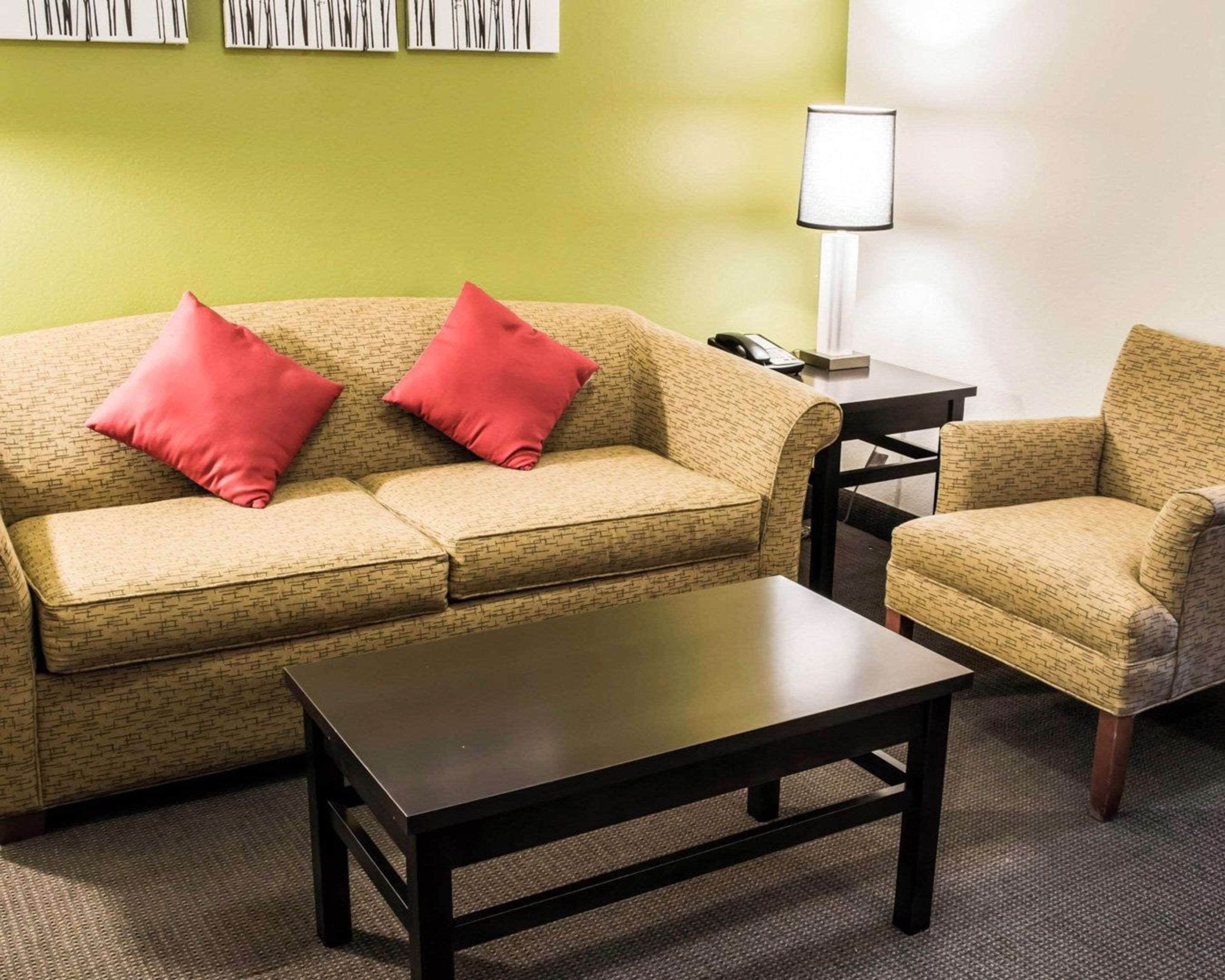 A living room with a beige patterned sofa, two red pillows, a black coffee table, a beige armchair, and a small side table with a lamp.