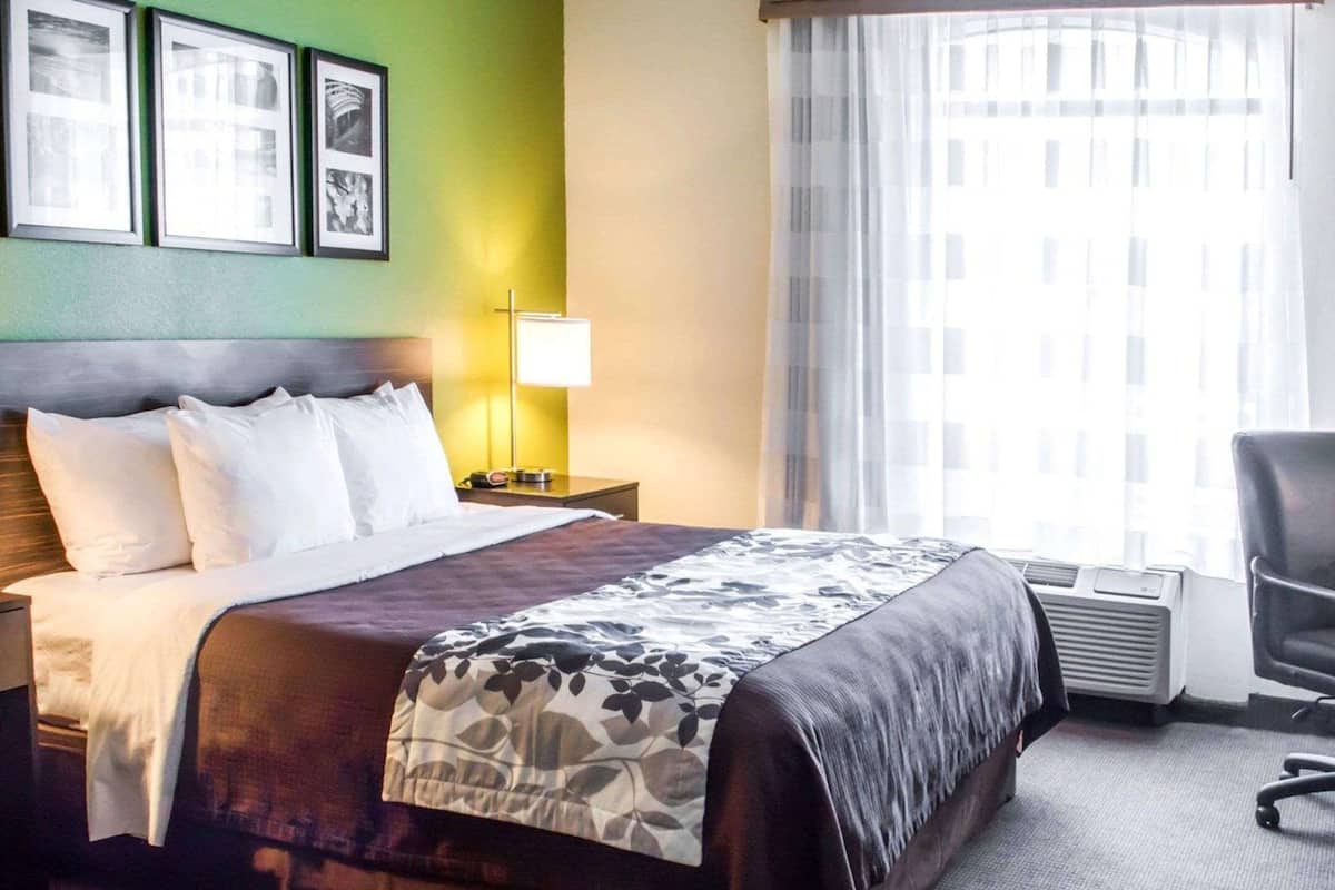 A hotel room with a bed, a nightstand, a lamp, a chair, and a window with curtains.