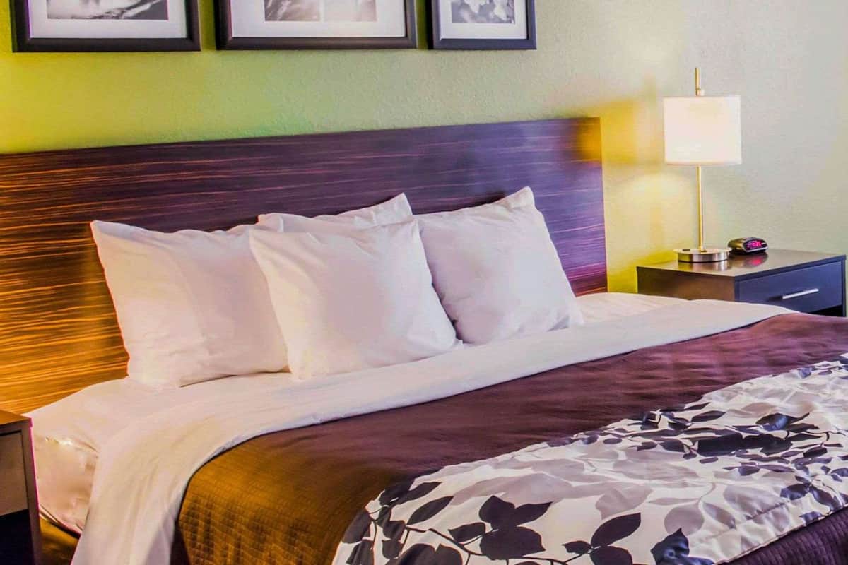 A neatly made bed with white linens and a floral-patterned throw, a dark wooden headboard, and a nightstand with a lamp and a clock.