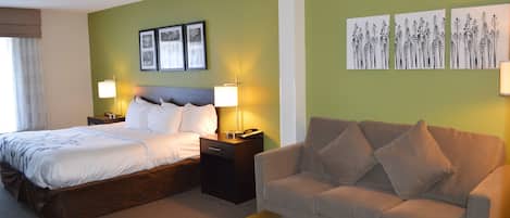 Quarto de hotel com cama, mesa de cabeceira, sofá e mesa de centro. Há luminárias fixadas na parede e quadros emoldurados.