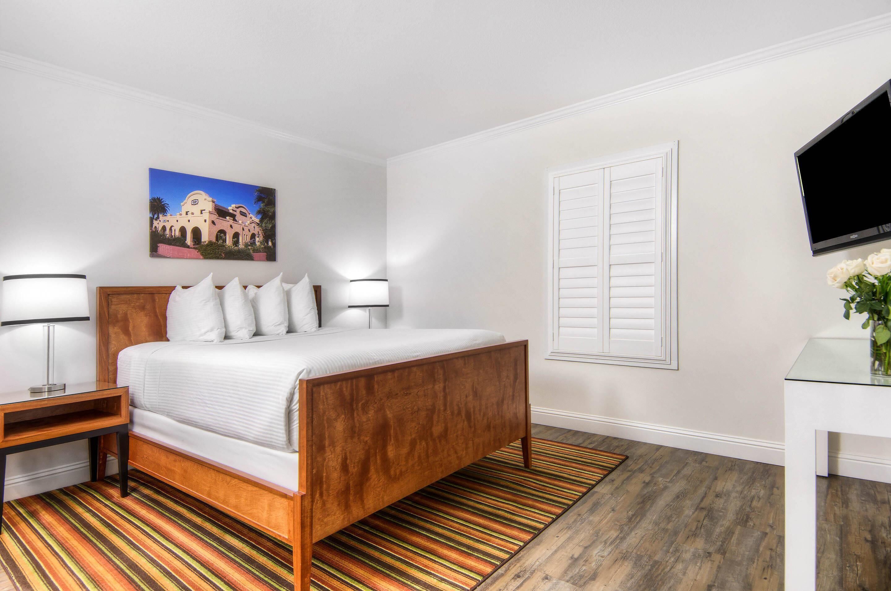 A hotel room with a wooden bed, white walls, a painting of a building, a TV, and a small table with a lamp.