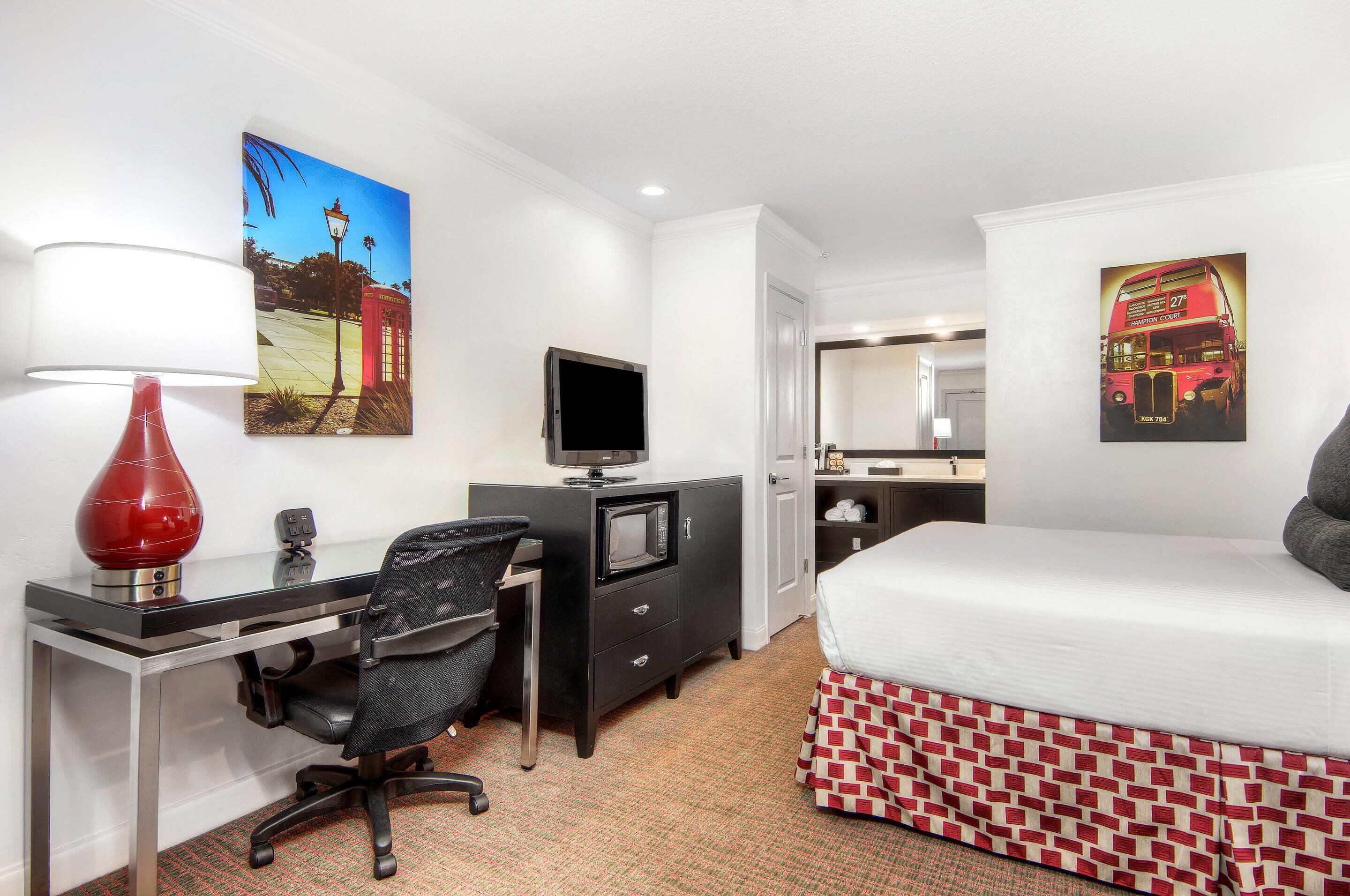 Habitación de hotel con cama, escritorio, silla, televisor y obras de arte en las paredes.