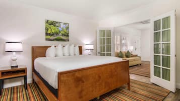 A hotel room with a wooden bed, white bedding, a nightstand, a lamp, a painting of a park, a sofa, and a door with green curtains.