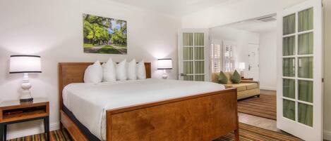 A hotel room with a wooden bed, white bedding, a nightstand, a lamp, a painting of a park, a sofa, and a door with green curtains.