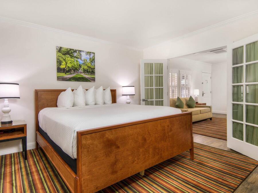 A hotel room with a wooden bed, white bedding, a nightstand, a lamp, a painting of a park, a sofa, and a door with green curtains.