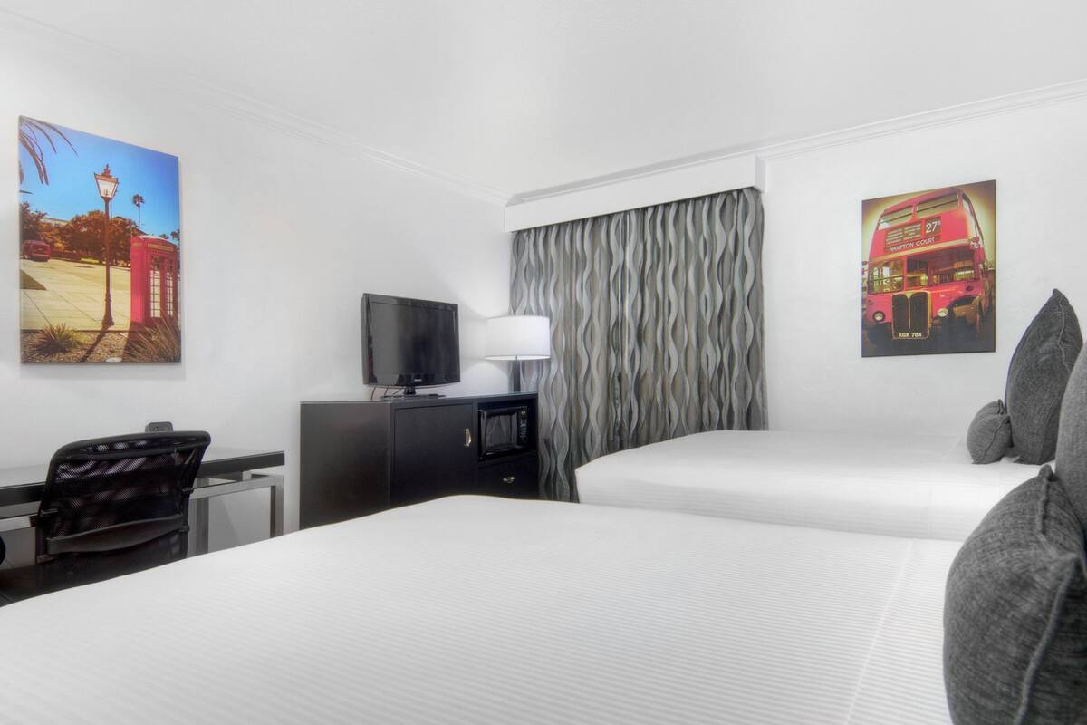 A hotel room with two beds, a TV, a desk with a chair, a microwave, and a window with curtains.