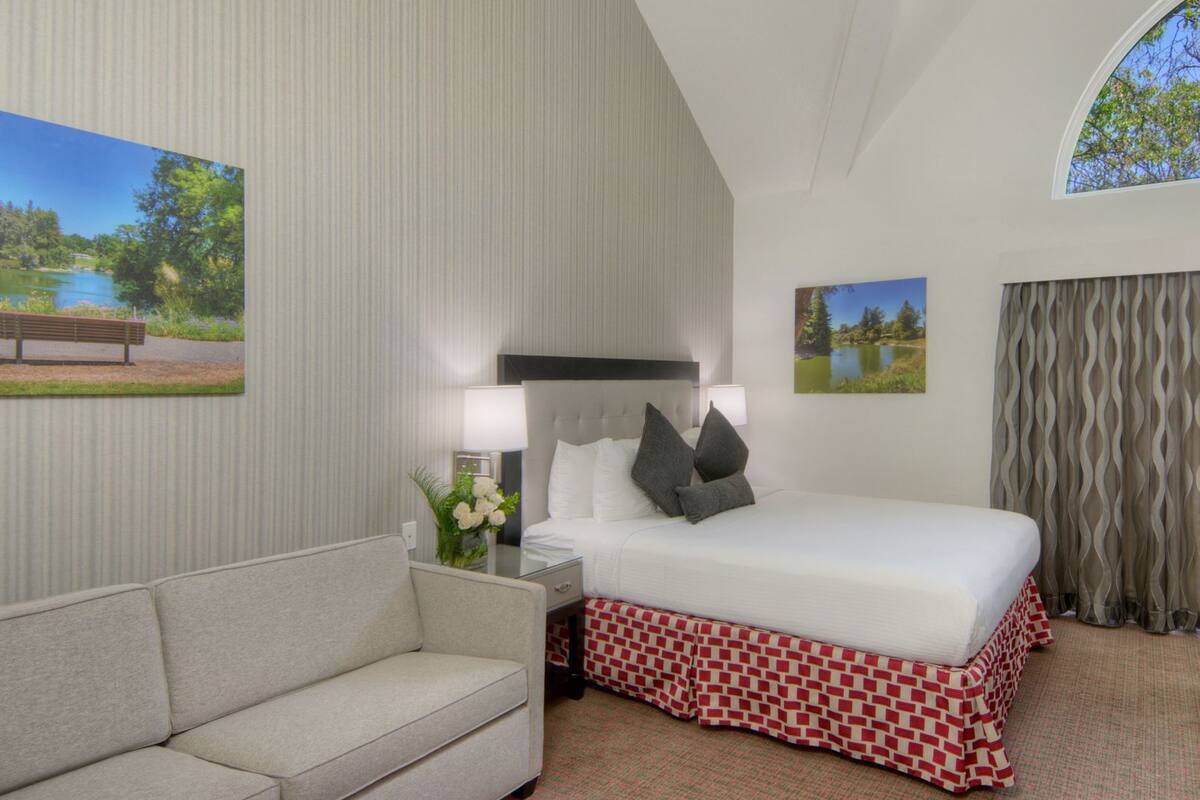 A hotel room with a bed, a sofa, a nightstand with a lamp, and two framed pictures on the walls.