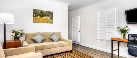 A living room with a beige sofa, a striped rug, a wooden side table with a vase of flowers, and a flat-screen TV mounted on the wall.