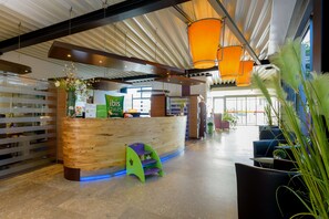 A hotel reception area with a wooden reception desk, a green and purple children's play area, and orange hanging lamps. - ibis Styles Leipzig (Schkeuditz)