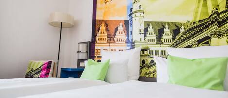 A hotel room with a bed, pillows, a lamp, and a wall with a cityscape poster.