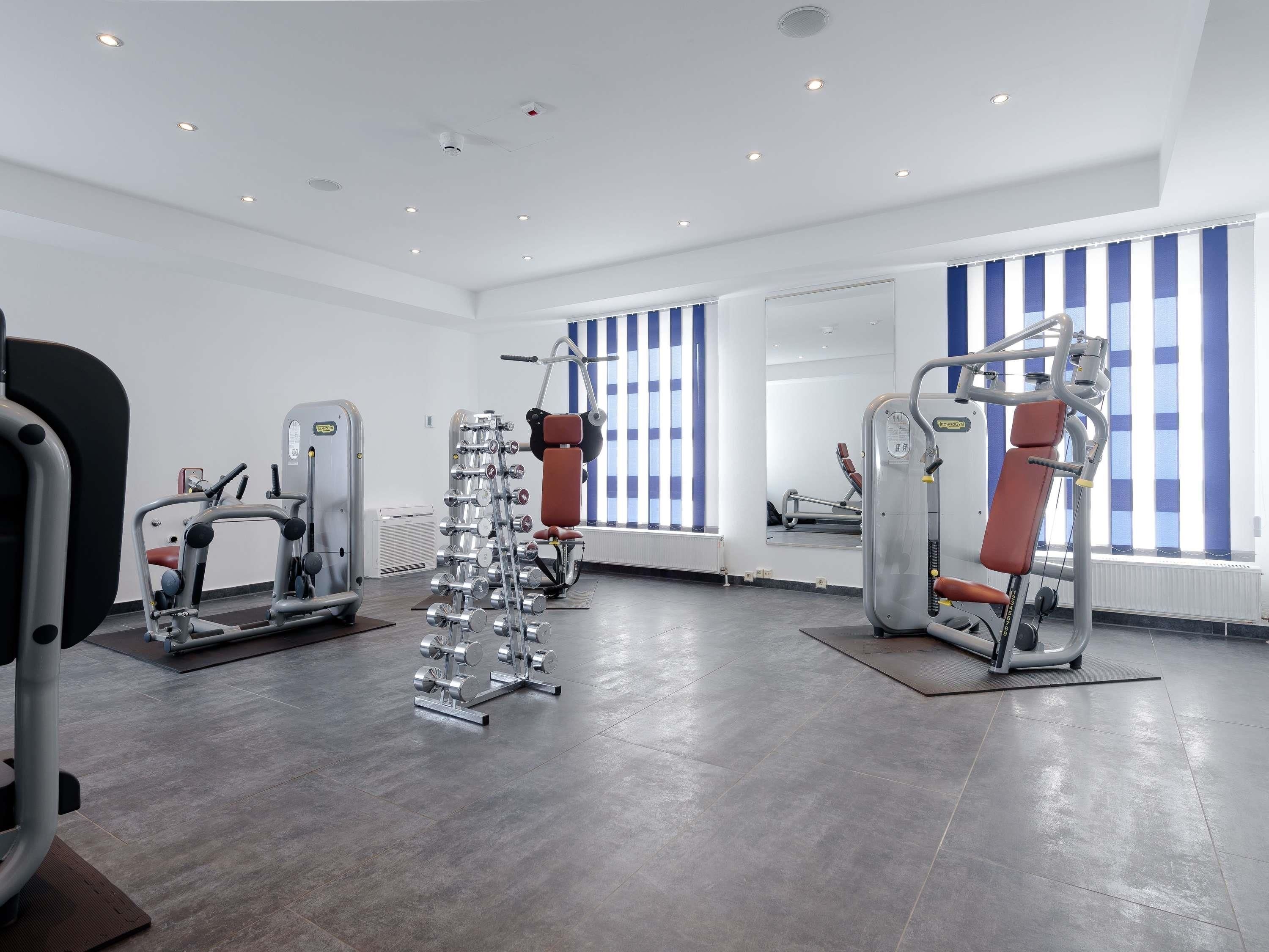 A modern gym with various exercise equipment, large windows with blue and white striped blinds, and a clean, spacious layout.