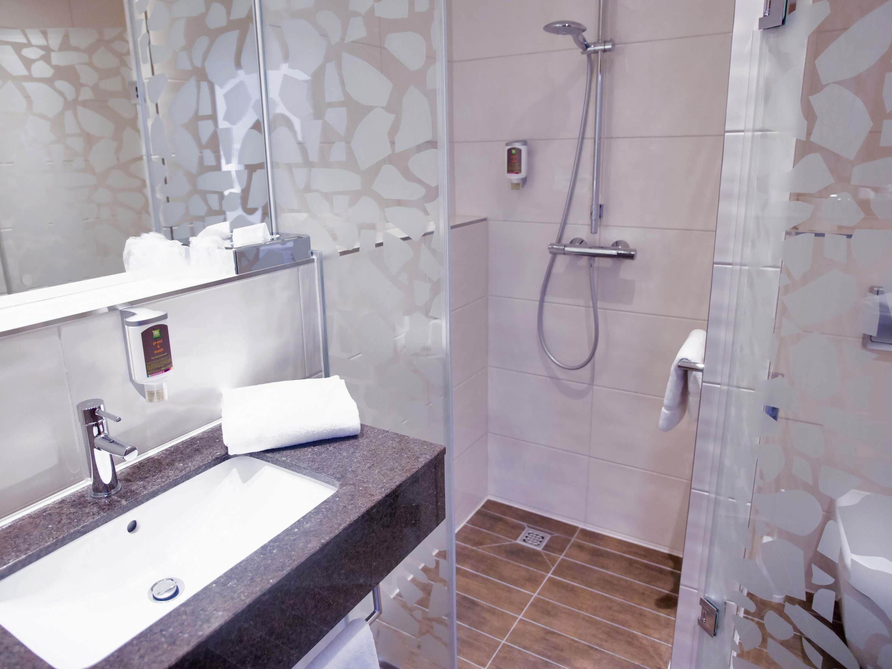 Un baño con lavamanos, espejo, toalla, ducha y zona de ducha con mampara de cristal.