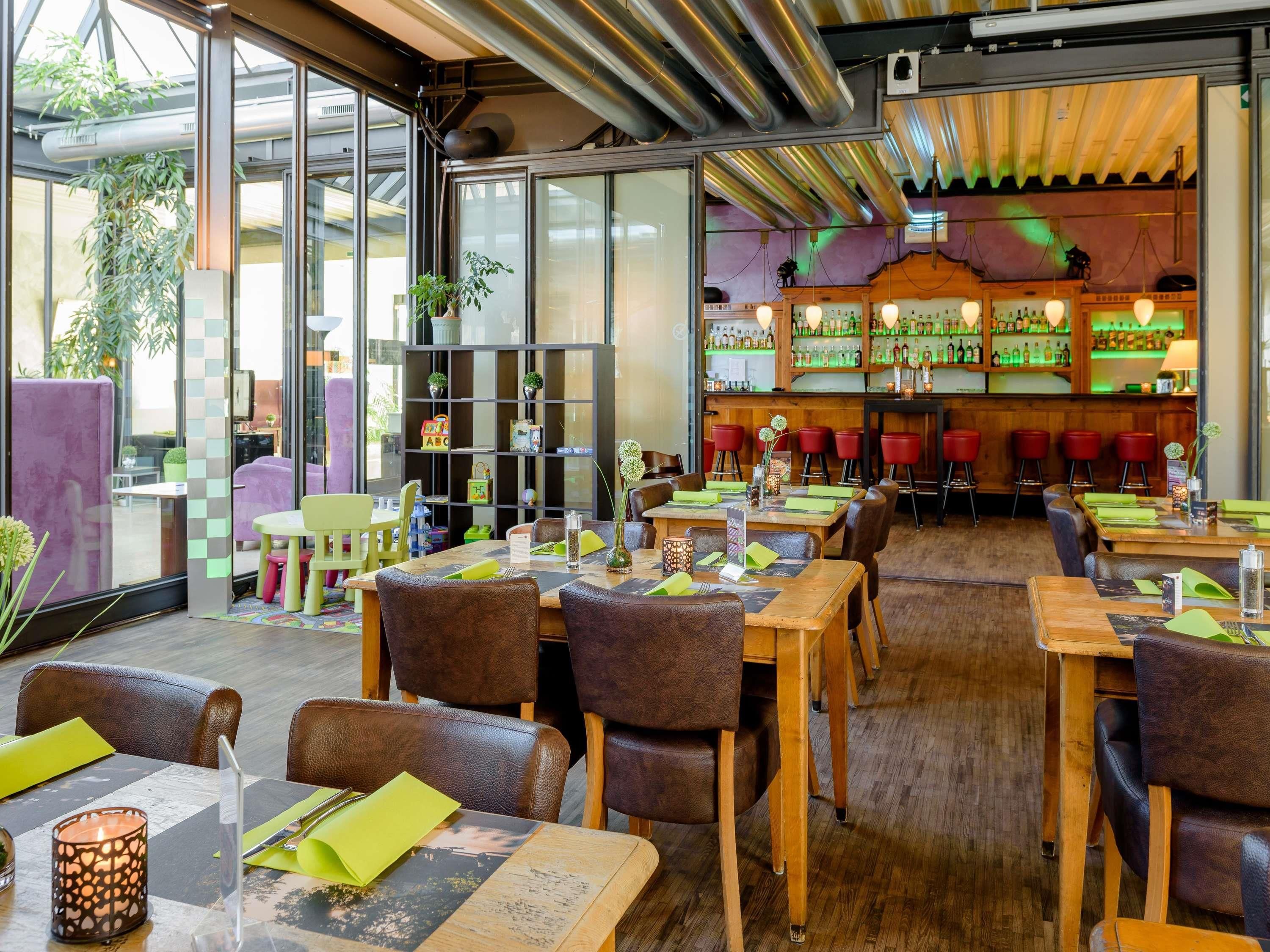 A restaurant with wooden tables and chairs, a bar area with bottles, and a large window with plants.