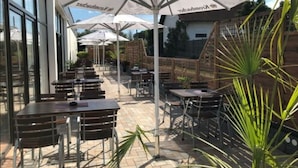 An outdoor patio area with multiple tables and chairs, covered by umbrellas, and surrounded by glass walls. - ibis Styles Leipzig (Schkeuditz)