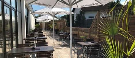 An outdoor patio area with multiple tables and chairs, covered by umbrellas, and surrounded by glass walls.