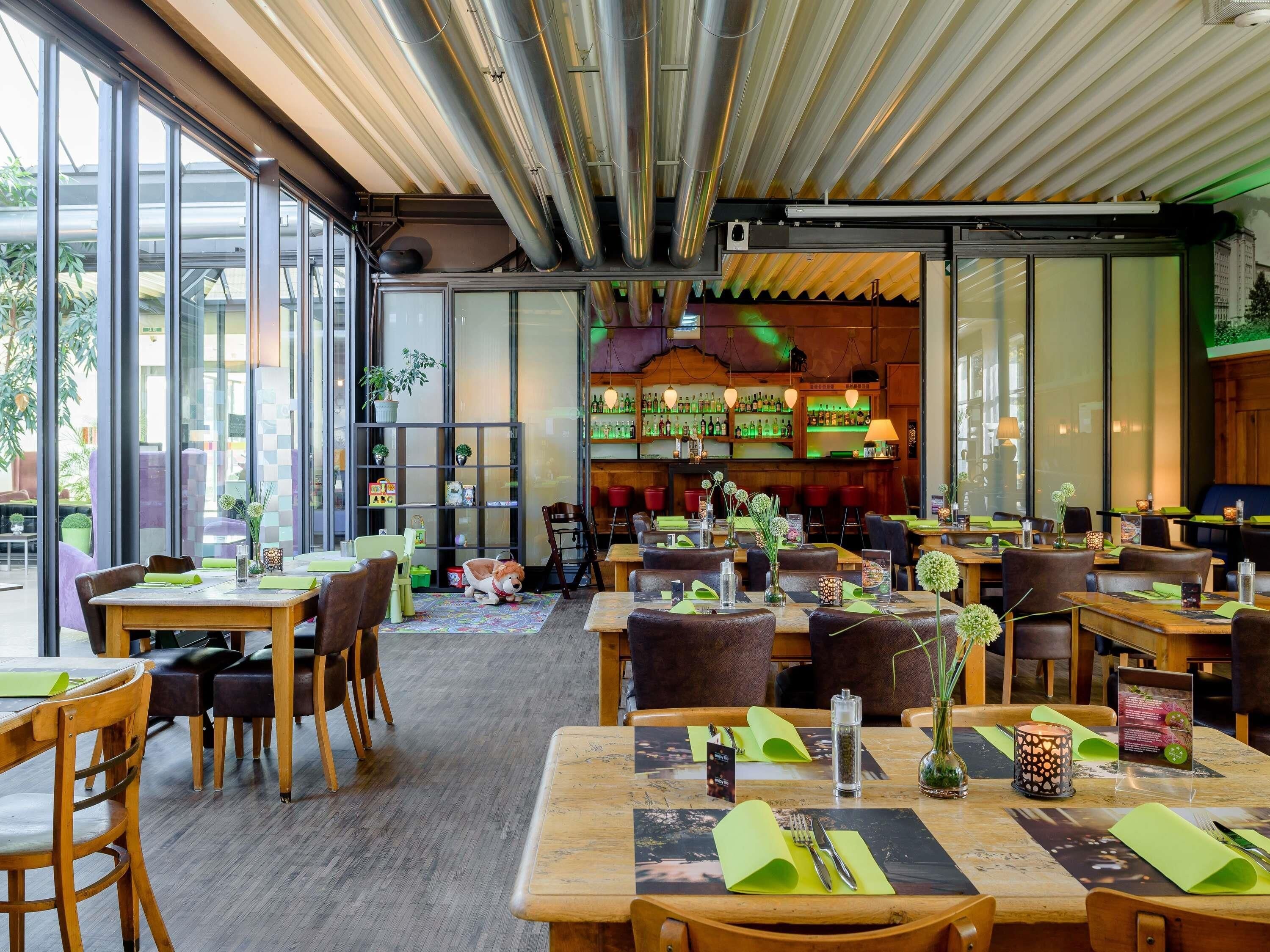 En restaurang med en modern inredning, med träbord och stolar, en bardisk med hyllor fulla av flaskor och stora fönster som släpper in naturligt ljus.