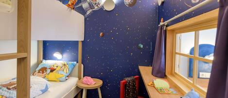 A child's room with bunk beds, a small stool, a red chair, a window with a view, and a wall with a space-themed mural.