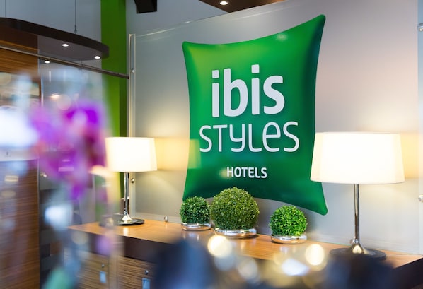 Un espace d’accueil avec une banderole verte sur laquelle on peut lire « ibis Styles Hotels » et deux lampes posées sur un comptoir en bois.