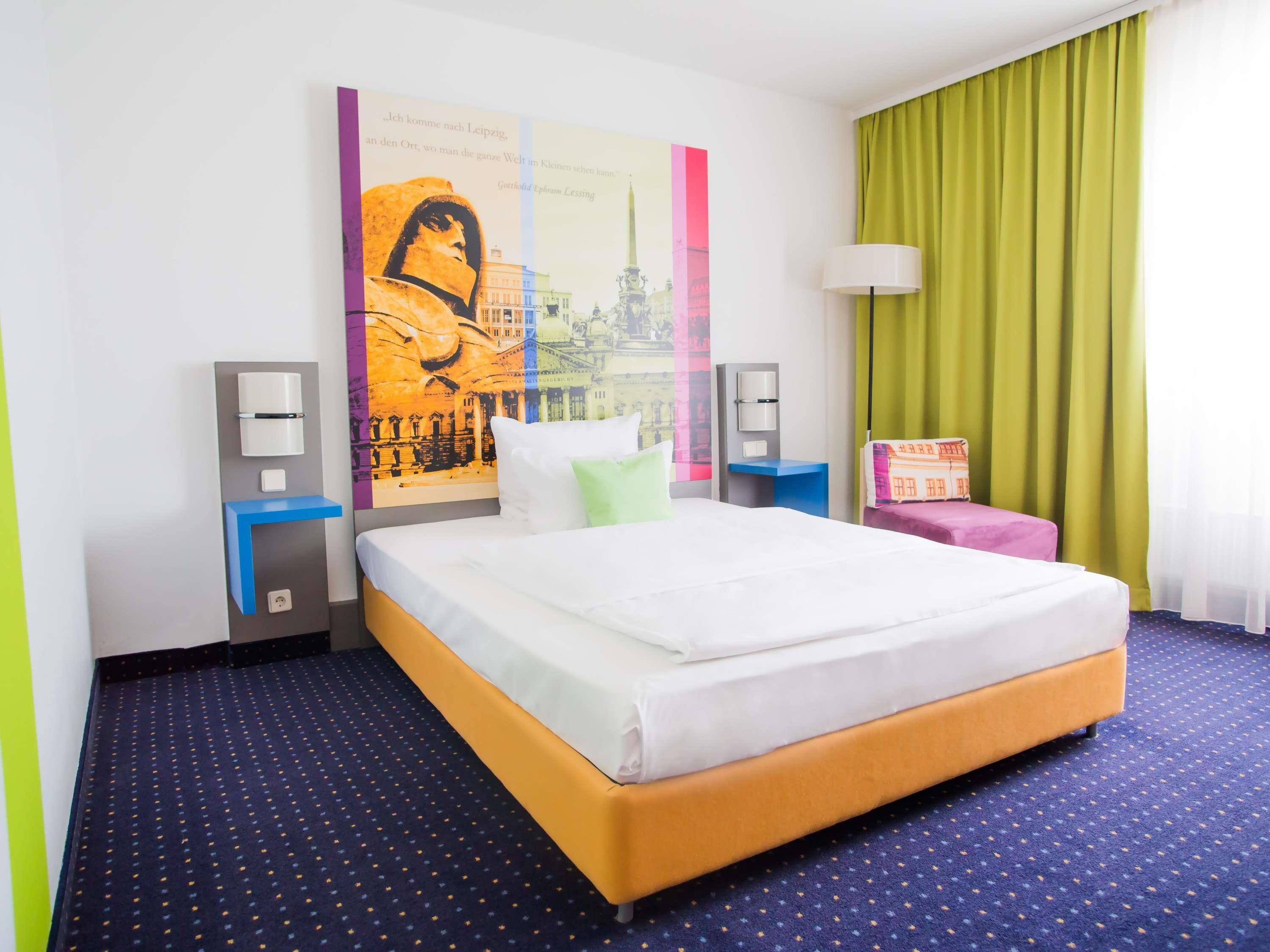 A hotel room with a large bed, a nightstand with a lamp, a wall-mounted speaker, and a poster of a cityscape.