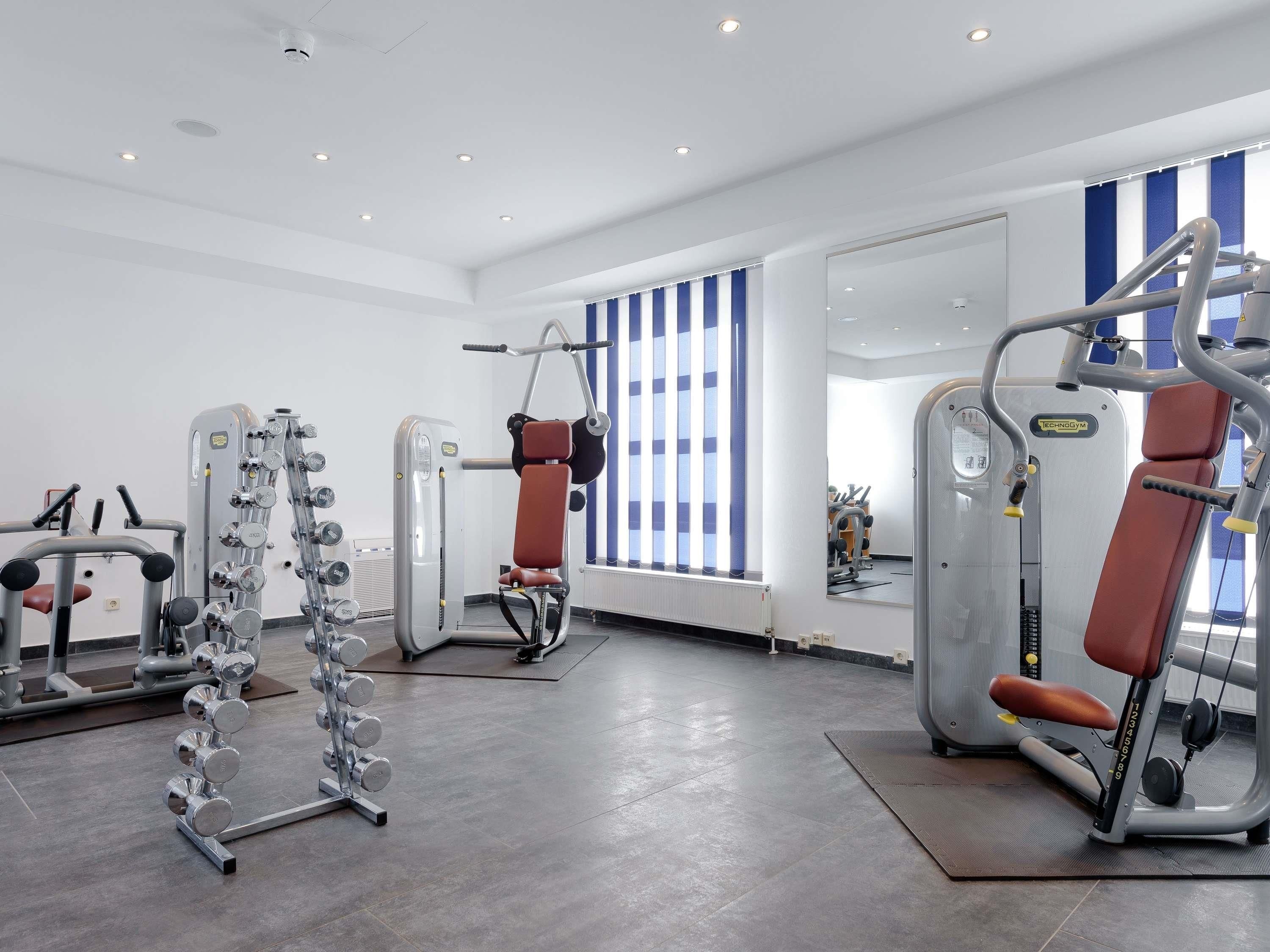 Une salle de sport équipée de divers appareils d’exercice, dotée d’un mur recouvert de miroirs et d’une fenêtre à rayures bleues et blanches.