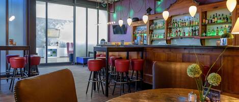 Una zona de bar con mesas de madera y taburetes rojos, una barra con botellas y un ventanal con vistas.