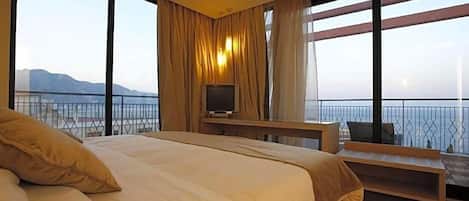 A bedroom with a bed, a TV, a desk, and large windows with a view of the sea.