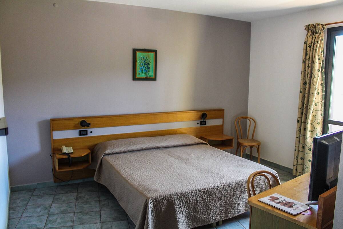 A hotel room with a bed, bedside table, chair, desk, television, and a framed picture on the wall.