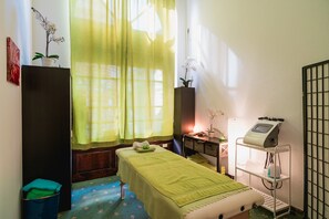 Una habitación con cama, mesa de masaje, un mueble pequeño, un cubo de basura y una ventana con cortinas translúcidas.