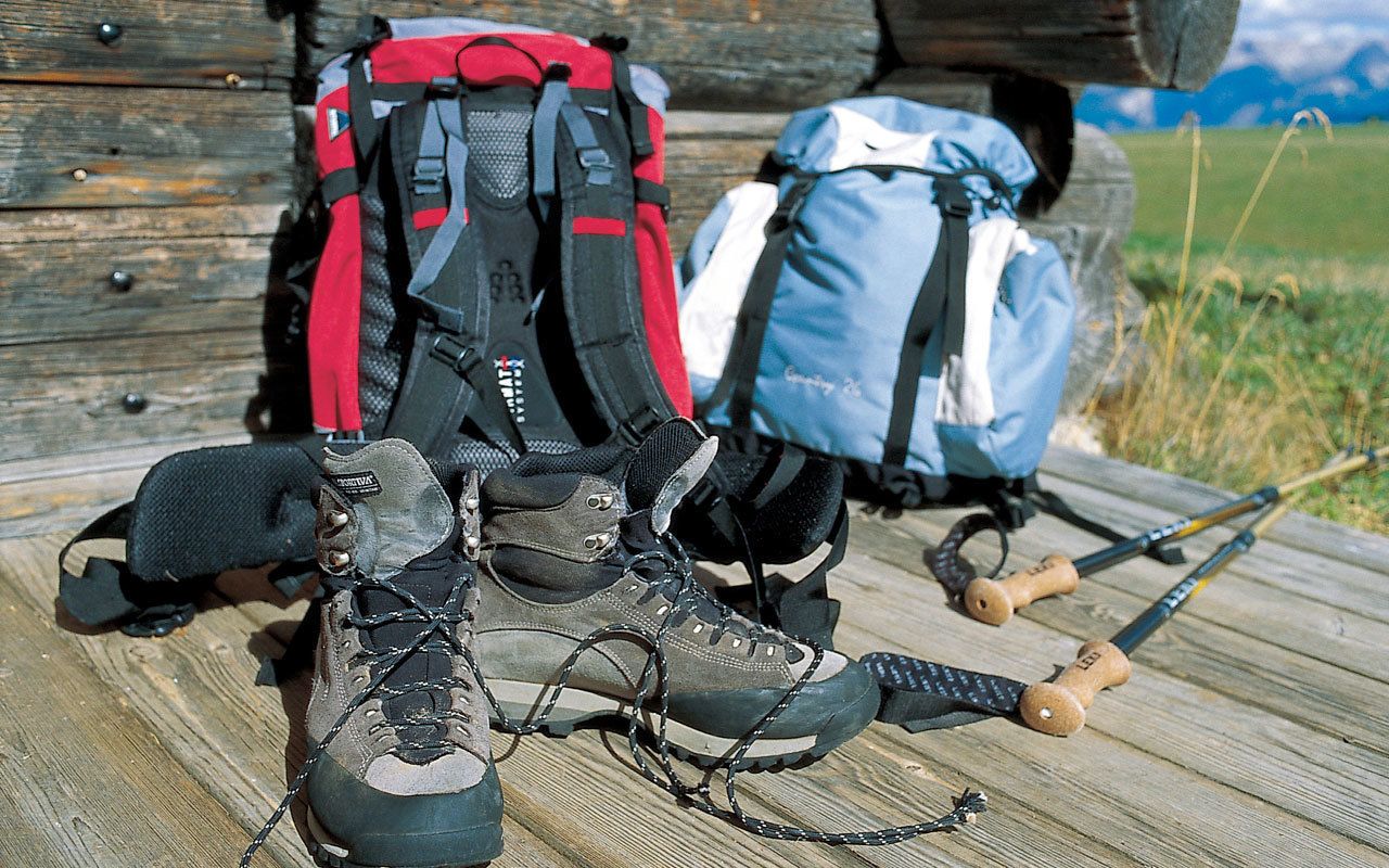 Un par de botas de senderismo, una mochila roja y negra, y una mochila azul sobre una superficie de madera con una estructura de madera al fondo.