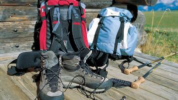 Un par de botas de senderismo, una mochila roja y negra, y una mochila azul sobre una superficie de madera con una estructura de madera al fondo.