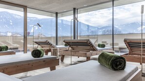 A spa room with wooden loungers, a massage table, and a view of snow-capped mountains through large windows. - Hotel Langgenhof (Brunico)