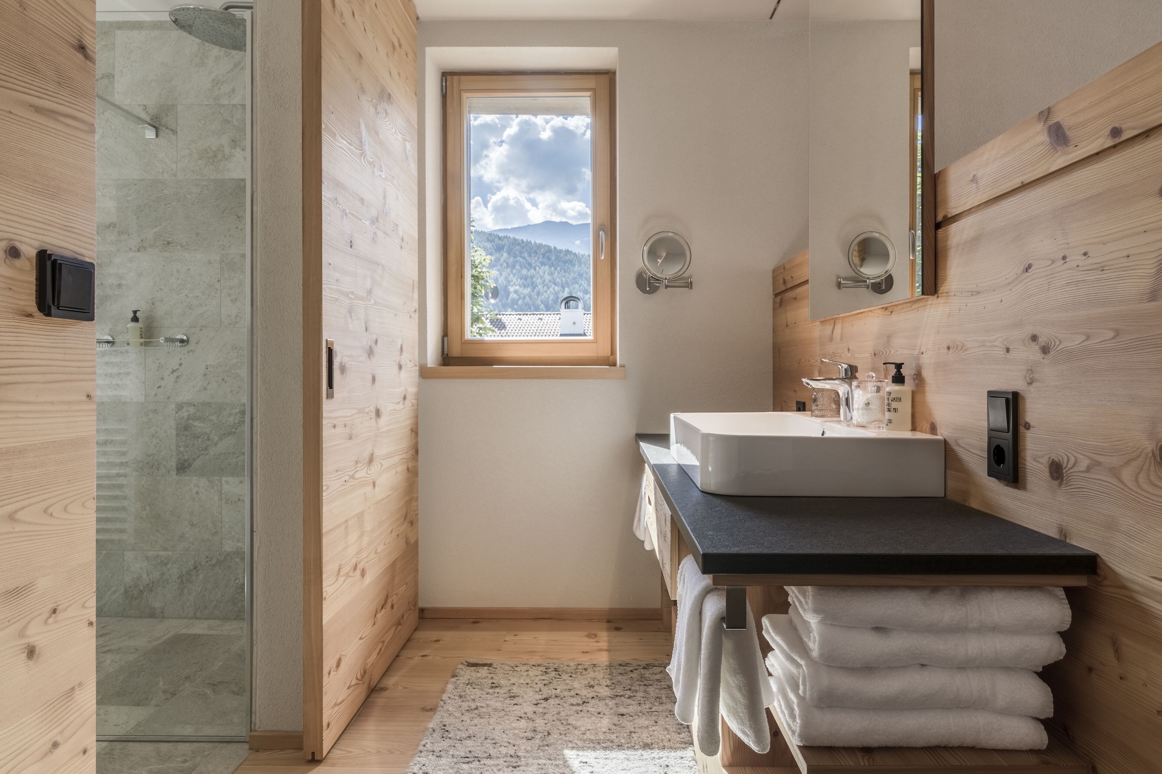 A modern bathroom with a wooden interior, a glass shower enclosure, a sink with a black countertop, and a window with a mountain view.