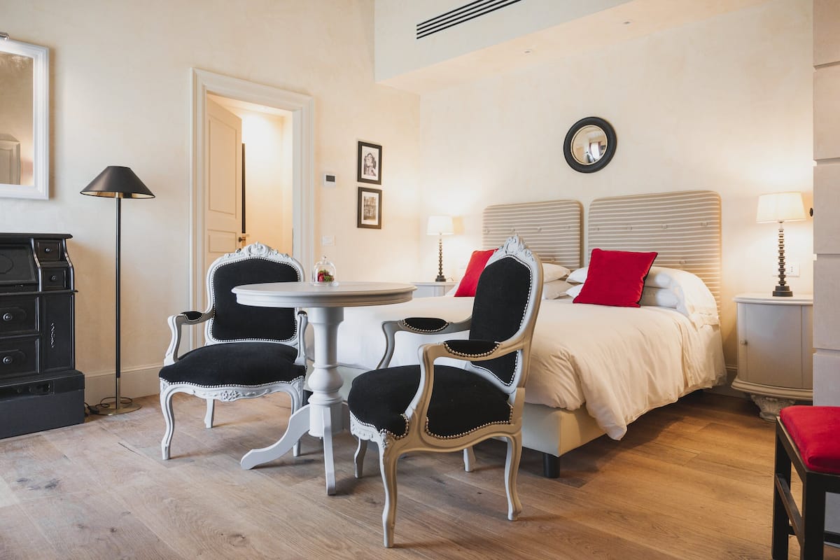 Un dormitorio con cama, dos sillas, una mesa redonda, un aparador y un espejo en la pared.
