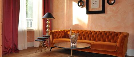 Ein Zimmer mit einem orangefarbenen, gepolsterten Sofa, einem runden Tisch mit einem Glas, einer Lampe und einem gerahmten Bild an der Wand.