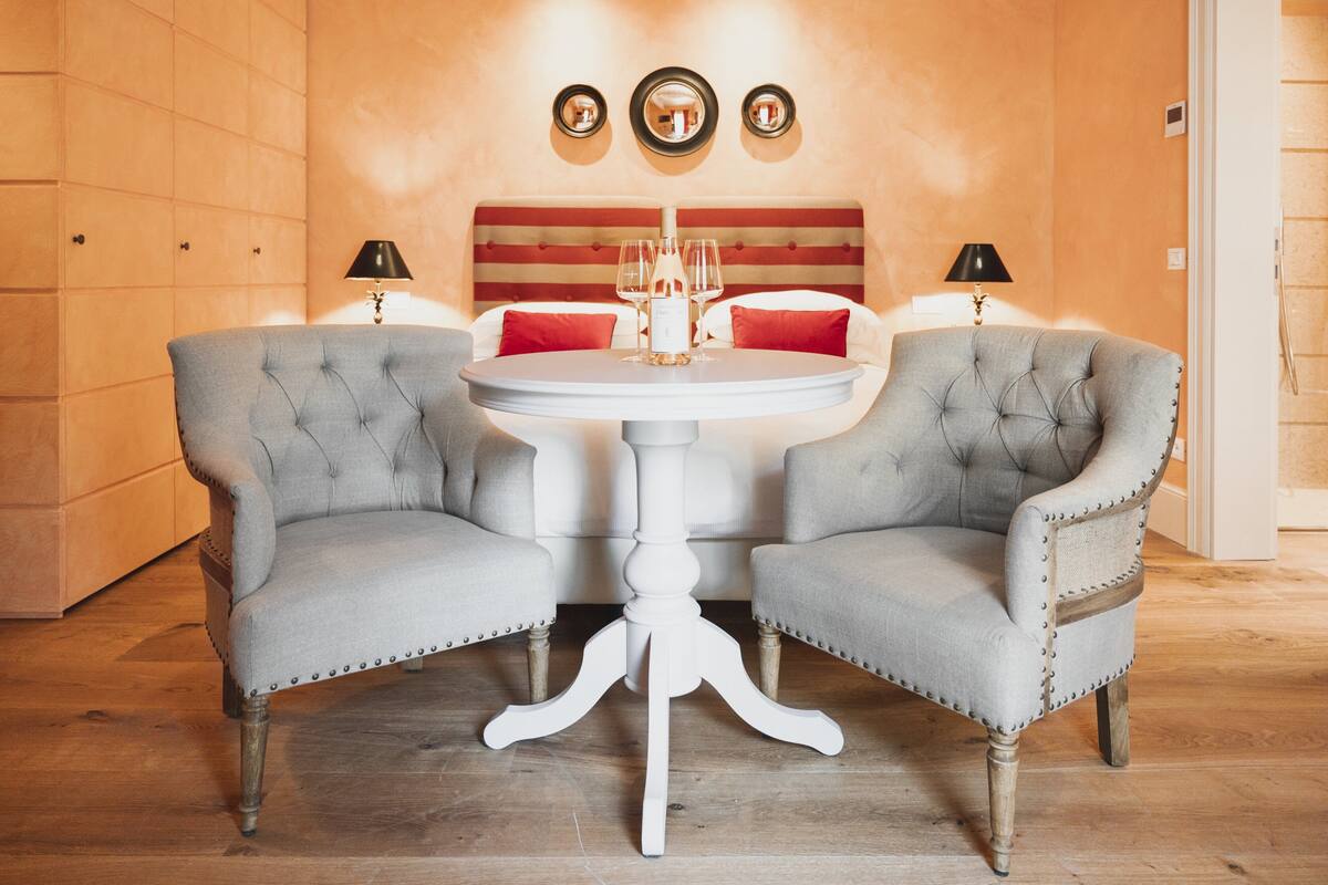 Una habitación con una mesa redonda blanca, dos sillas grises tapizadas, un cabecero rojo y blanco, y tres espejos fijos a la pared.