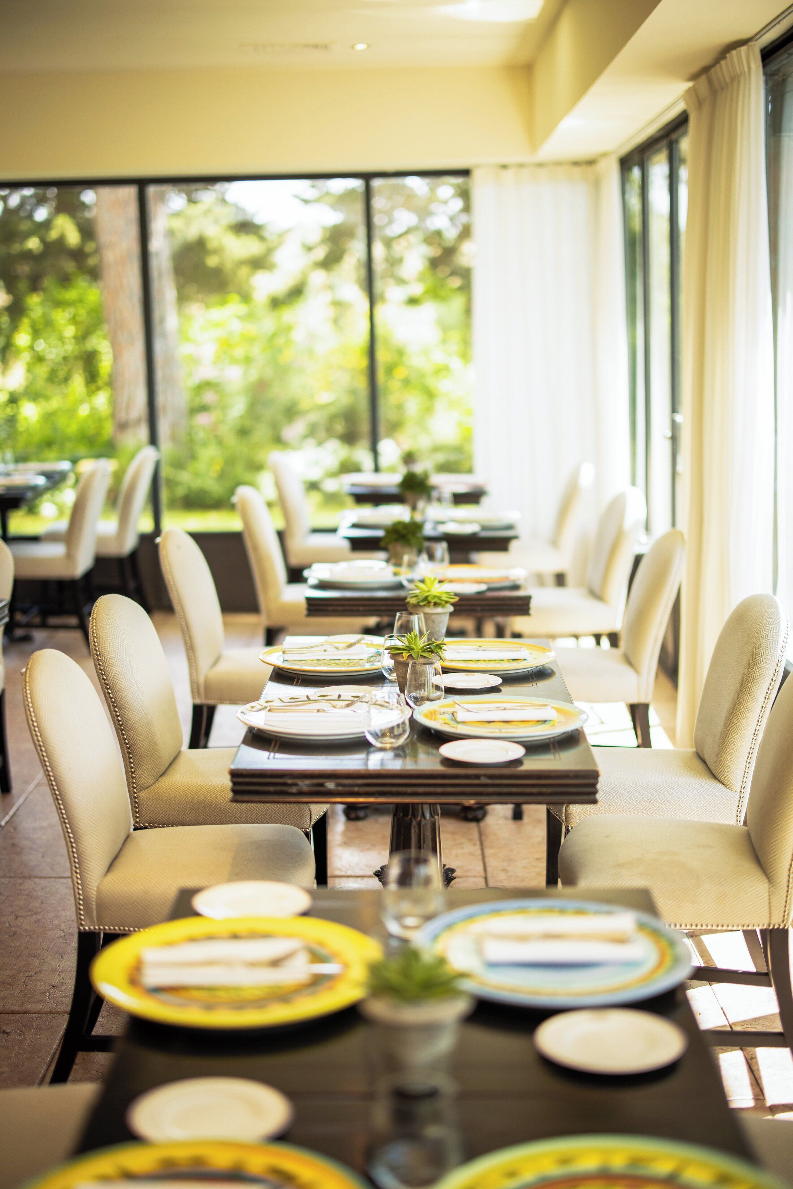 A dining area with tables set for a meal, chairs, and a view of greenery outside.