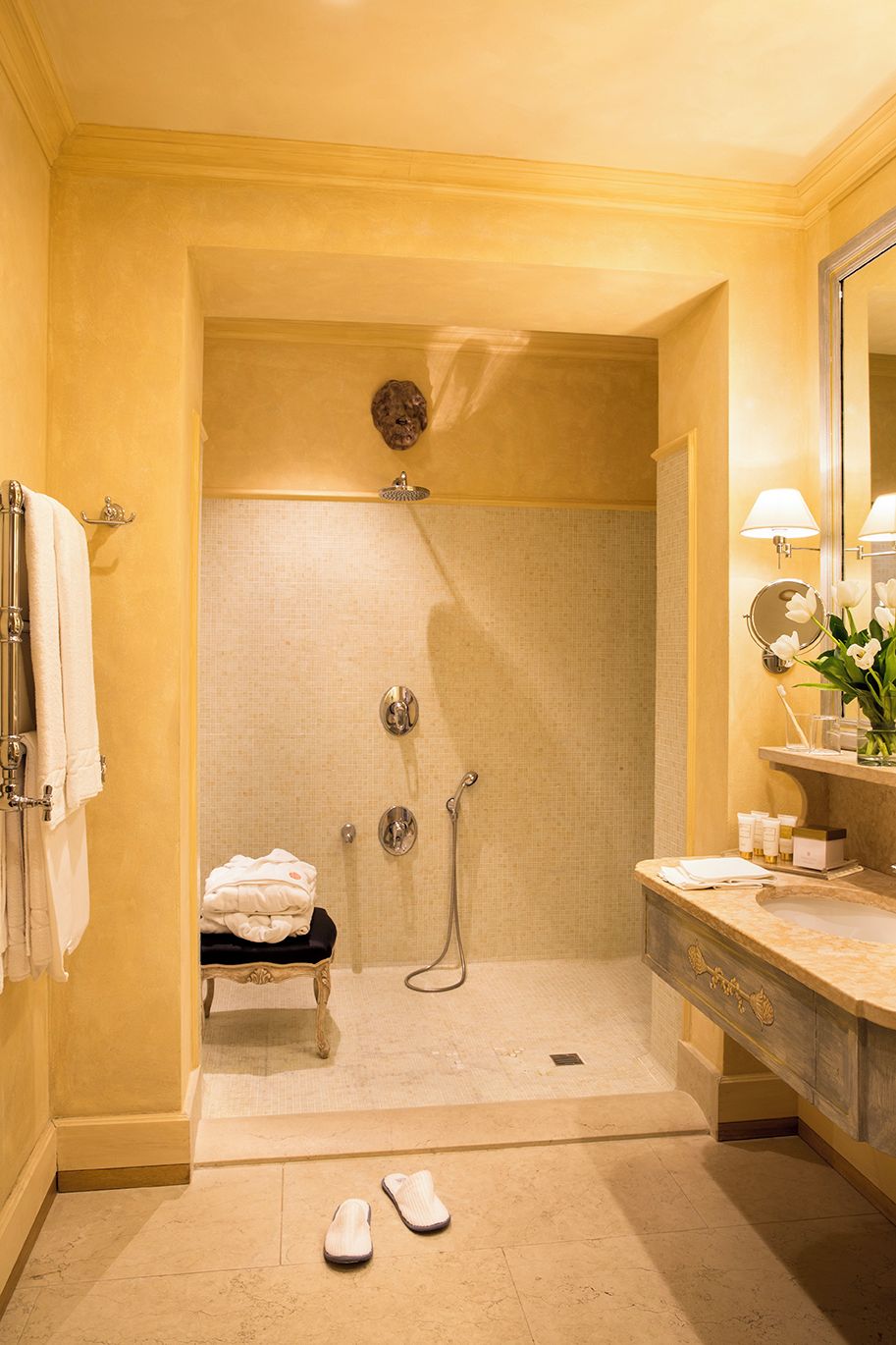 Un baño con ducha, lavamanos y espejo. Hay toallas y zapatillas en el suelo.