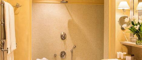 Un baño con ducha, lavamanos y espejo. Hay toallas y zapatillas en el suelo.