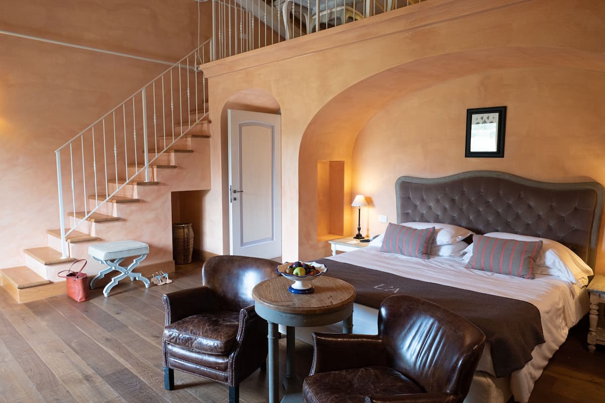 Habitación de hotel con una cama grande, una mesa redonda con fruta, sillas de cuero, una escalera y un cuadro en la pared.