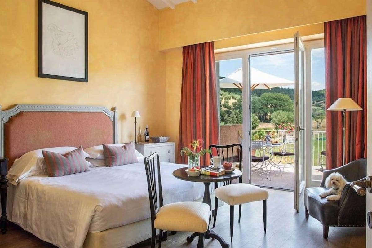Una habitación de hotel con cama, una mesita con un florero, una silla, un balcón con mesa y sillas, y un perro en una silla.