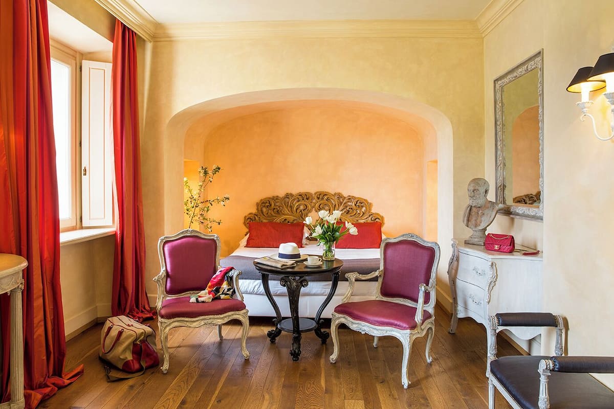 Una habitación de hotel con una cama, dos sillones, una mesa con un florero, un busto en un tocador y un espejo en la pared.