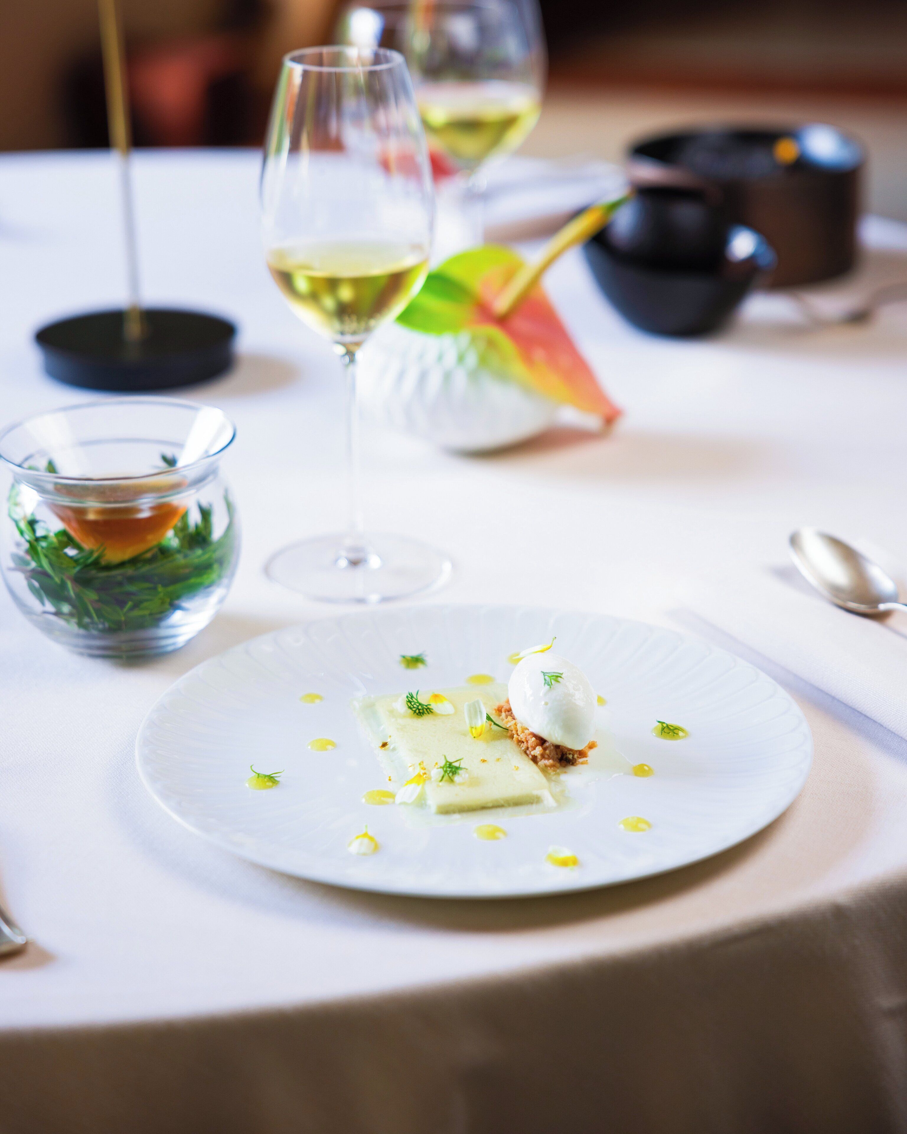 A dining table with a white tablecloth, a glass of white wine, a small bowl of green garnish, and a white plate with a dessert.