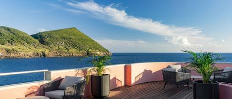 A deck with outdoor seating, potted plants, and a view of the sea and hills.